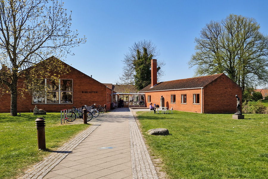 Flisebelagt sti med græs på begge sider, der fører hen til Nyborg Bibliotek i baggrunden. Rød murstensbgning med tegltag.